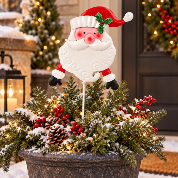 A decorative Santa Claus yard stake with a smiling white beard, red hat, and green holly accent, displayed in a snowy evergreen planter with pinecones and red berries in front of a holiday door.