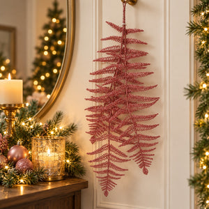 A pink glittery artificial fern hanging on a white paneled door in a decorated holiday setting with a mirror, candle, and warm lights.