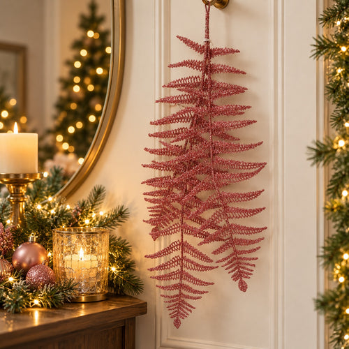 A pink glittery artificial fern hanging on a white paneled door in a decorated holiday setting with a mirror, candle, and warm lights.