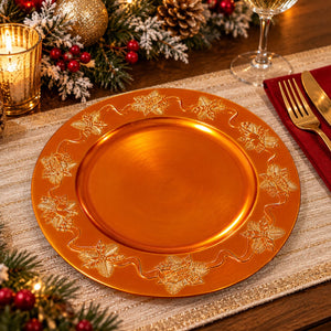 A round orange-gold charger plate with a raised gold floral border, styled on a table runner with a candle, wine glass, fork, knife, and holiday greenery in the background.