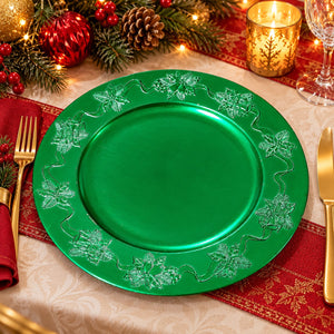 A green round charger plate with embossed holly and vine patterns, styled on a holiday table setting with a gold fork, gold knife, red napkin, red placemat, pinecones, and a lit candle.