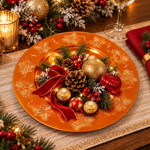 A festive Christmas table setting featuring a large copper-orange charger plate with embossed gold snowflakes, decorated with pine branches, red berries, pinecones, gold and red ornaments, a red ribbon, and Ferrero Rocher chocolates. A lit candle, wine glass, and red napkin are visible in the background.