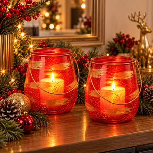 Two red glittery glass lanterns with gold wire handles, each containing a lit white candle, displayed on a wooden table surrounded by Christmas decorations including pine branches, red berries, pinecones, and warm string lights.