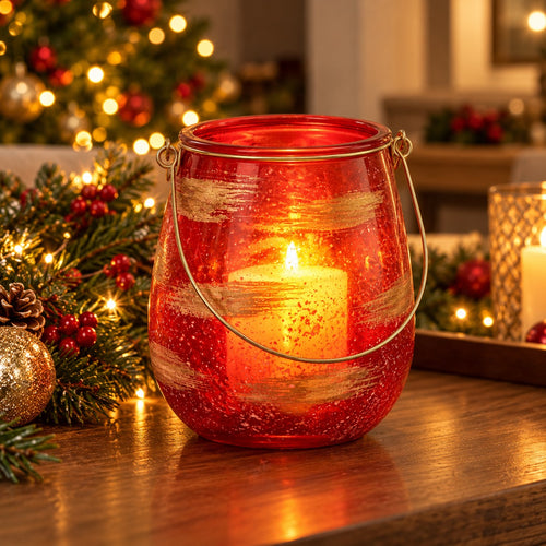 A red glass lantern candle holder with a wire handle sits on a wooden table, lit with a warm glowing candle inside. Christmas greenery with red berries and warm string lights are visible in the softly blurred background.