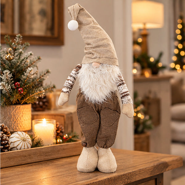 A rustic Christmas gnome figurine standing on a wooden table in a warmly lit holiday room. The gnome has a tall beige hat with a white pom-pom, a fluffy white beard, brown pants, and cream-colored shoes.