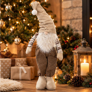 A Christmas gnome decoration standing on a wooden floor in a cozy holiday setting, with a lit Christmas tree, wrapped gifts, pinecones, and a lantern in the background.