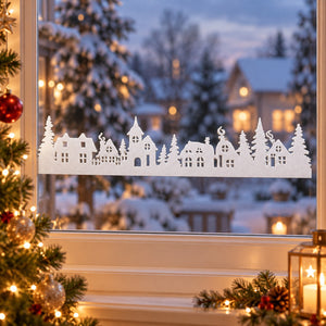 A white paper cutout of a Christmas village with houses and trees, displayed on a window during a snowy evening. The window frame is decorated with warm lights, pine branches, and candles, and a decorated Christmas tree is visible on the left.