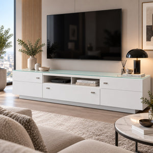 A modern white TV console with a translucent green-tinted glass top, styled in a contemporary living room with a wall-mounted black television, plants, and a round marble coffee table in the foreground.