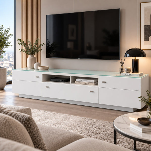A modern white TV console with a translucent green-tinted glass top, styled in a contemporary living room with a wall-mounted black television, plants, and a round marble coffee table in the foreground.