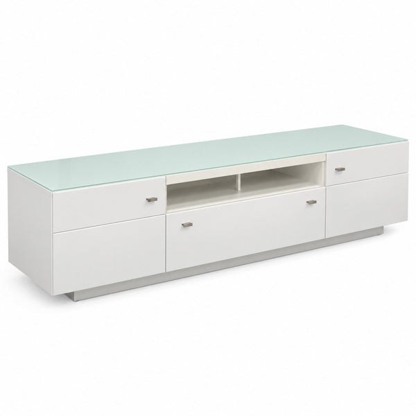 A modern white media console with a light green glass top, featuring three drawers, a central open shelf, and two side drawers with small silver handles.