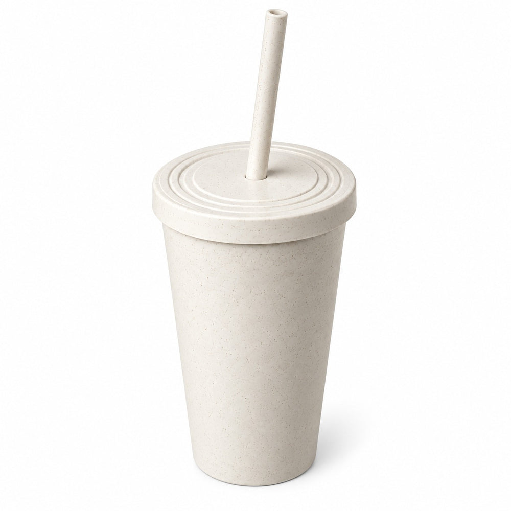 A light beige textured reusable tumbler with a matching lid and straw, standing on a wooden desk in a bright home office setting with a laptop, books, and a plant in the background.