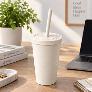 A light beige textured reusable tumbler with a matching lid and straw, standing on a wooden desk in a bright home office setting with a laptop, books, and a plant in the background.