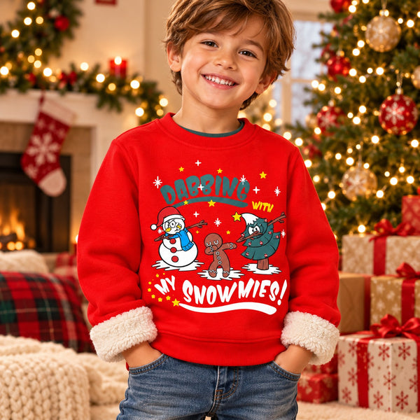 A young boy smiling in a cozy holiday living room, wearing a bright red Christmas sweatshirt with 'Carnival with my Snowmies!' printed on the front. The sweatshirt features a cartoon snowman, gingerbread man, and Santa, and has white faux-fur trimmed cuffs.