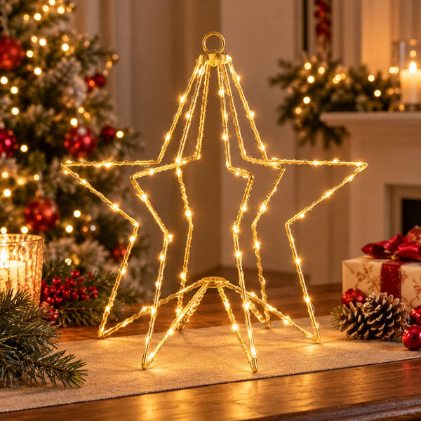 A large star-shaped wire light frame glowing with warm yellow Christmas string lights, displayed on a table in a decorated holiday living room with a Christmas tree, candles, pinecones, and wrapped gifts in the background.