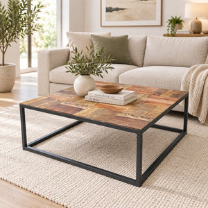 A square coffee table with a rustic multi-toned wood top and a black metal frame, styled in a bright living room next to a beige sofa.