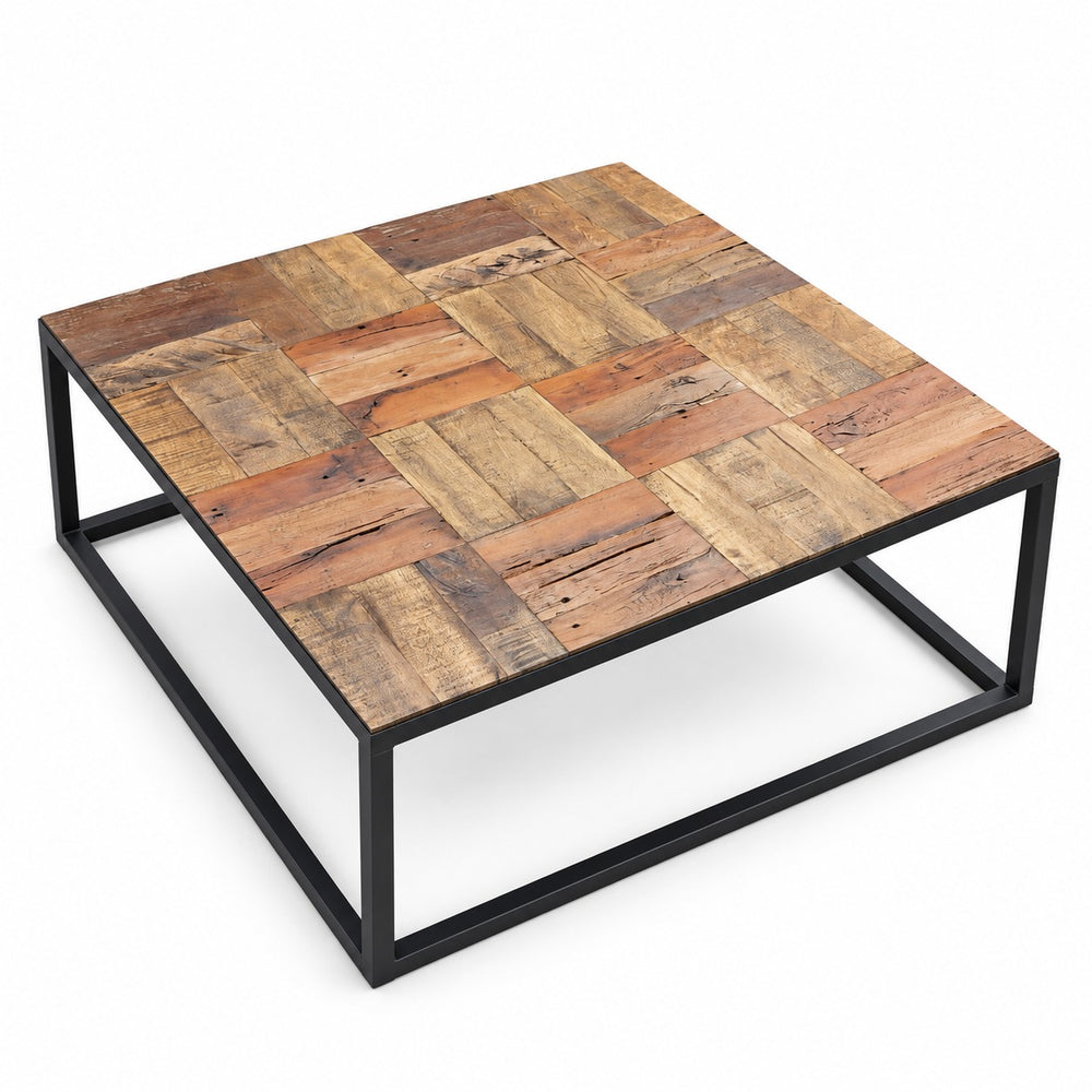 A square coffee table with a rustic patchwork wood top and a black metal frame, styled in a cozy living room next to a brown leather sofa.