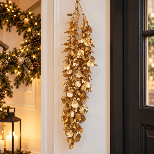 A hanging gold leaf garland decor piece displayed on a white wall next to a black door, with a softly lit holiday wreath and candles visible in the background.