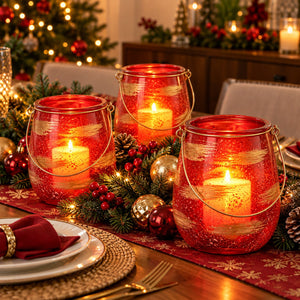 Three red glittery glass lanterns with metal handles, each containing a lit white pillar candle, arranged on a decorated Christmas table runner with pine branches, red berries, gold ornaments, and plates.