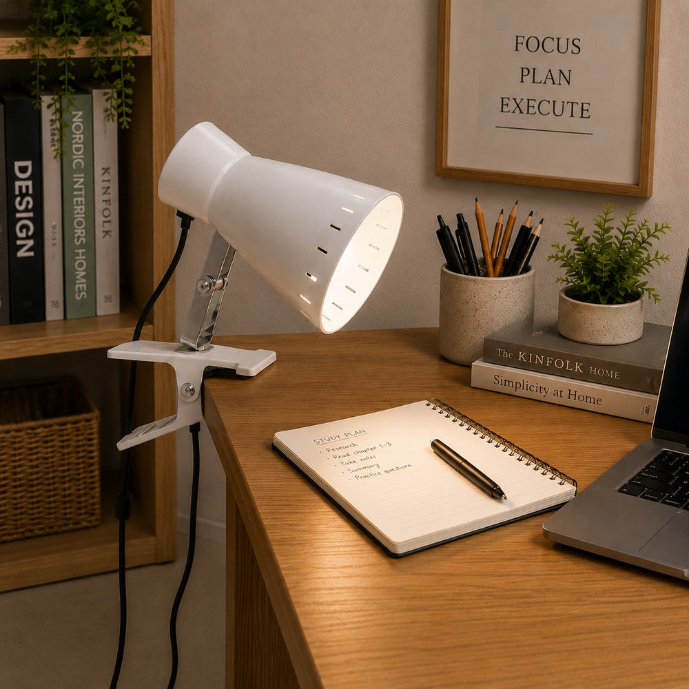 A white adjustable desk lamp with a clip base sits on a wooden desk in a styled home office setting, next to a notebook, laptop, books, and small plants.