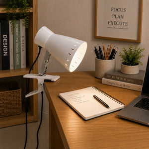 A white adjustable desk lamp with a clip base sits on a wooden desk in a styled home office setting, next to a notebook, laptop, books, and small plants.