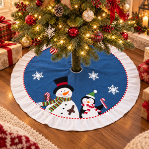 A round Christmas tree skirt with a blue center and white ruffled border, featuring cartoon snowman and penguin illustrations, shown under a decorated Christmas tree on a wooden floor.