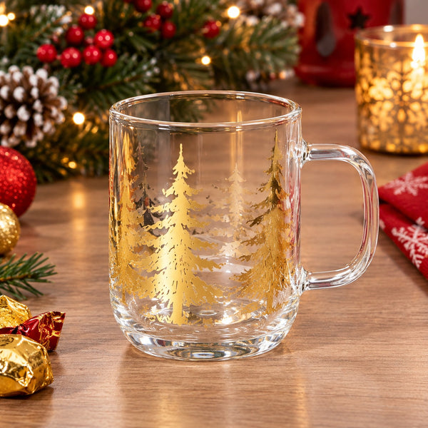 A clear glass mug with a gold foil tree design, set on a wooden table surrounded by festive Christmas decorations including a lit candle, pine branches, and red ornaments.