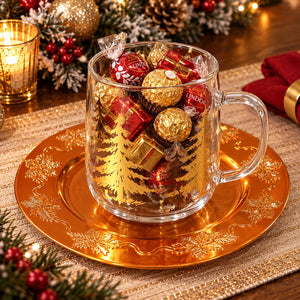 A clear glass mug filled with assorted wrapped chocolates, including Lindt and Ferrero Rocher, displayed on a gold-toned plate in a festive holiday setting with candles and pine branches.