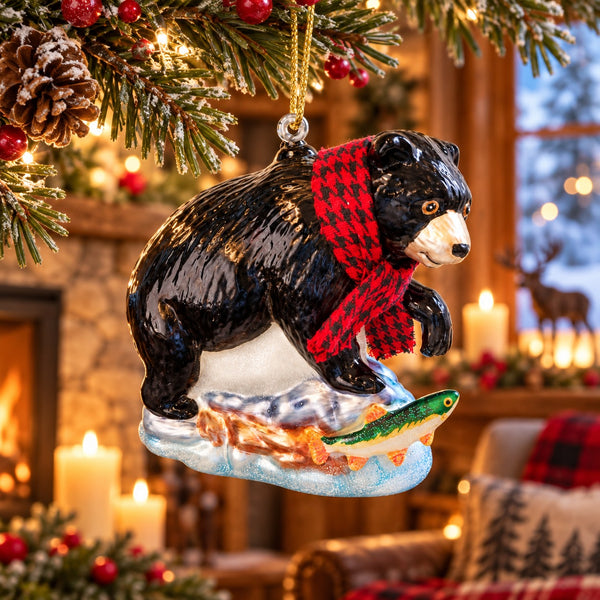 A Christmas ornament shaped like a black bear wearing a red and black plaid scarf and standing on a small boat with a green fish, hanging from a decorated pine branch in a warm, festive indoor setting.