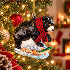 A Christmas ornament shaped like a black bear wearing a red and black checkered scarf, standing on a tree branch with a fish on its back. The ornament is hanging in front of a softly blurred Christmas tree and fireplace background.