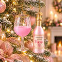 A Christmas ornament shaped like a pink wine bottle hanging on a decorated Christmas tree next to an ornament shaped like a pink wine glass. The bottle ornament is labeled 'Christmas Pink Rose' with a gold cap and gold hanging chain, set against a warm, softly blurred holiday background with lights and candles.