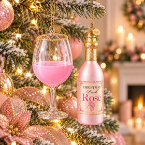 A Christmas ornament shaped like a pink wine bottle hanging on a decorated Christmas tree next to an ornament shaped like a pink wine glass. The bottle ornament is labeled 'Christmas Pink Rose' with a gold cap and gold hanging chain, set against a warm, softly blurred holiday background with lights and candles.