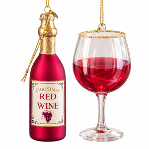 A Christmas ornament shaped like a bottle of red wine hanging beside a clear wine glass filled with red wine, on a plain white background.
