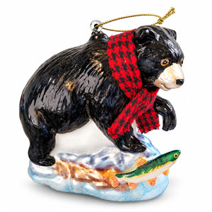A glossy black bear Christmas ornament wearing a red and black checkered scarf, standing on a snowy base with a green and orange fish.