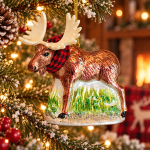 A ceramic Christmas ornament shaped like a moose standing on a grassy hill, hanging on a decorated Christmas tree. The ornament has a brown body, cream antlers and belly, a red-and-black checkered scarf, and a green-and-white base, with warm holiday lights in the background.