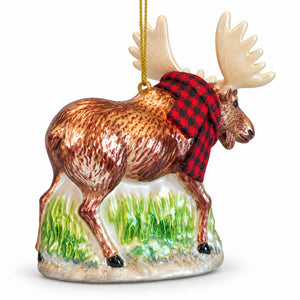A ceramic Christmas ornament shaped like a moose wearing a red and black plaid scarf, standing on a patch of green grass. The ornament has a gold hanging string and is photographed against a clean white background.