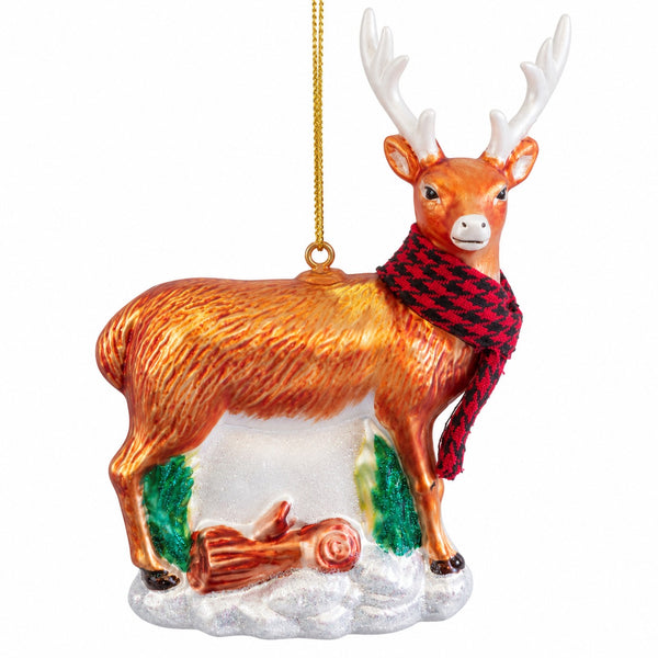 A decorative Christmas ornament shaped like a stag or deer with white antlers, wearing a red and black checkered scarf, standing on a snowy base with small green trees and a log.