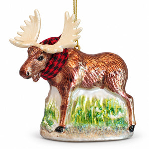 A ceramic moose ornament with large cream antlers, a brown body, a red and black plaid scarf, and a green grassy base, hanging by a gold string on a white background.