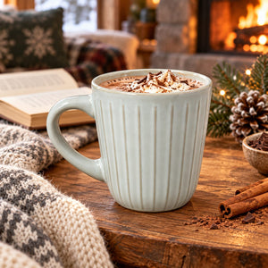 A ribbed ceramic mug filled with a hot cocoa drink topped with whipped cream and cocoa powder, set on a wooden table in a cozy indoor scene with a knit blanket, cinnamon sticks, pinecones, and a fireplace in the background.