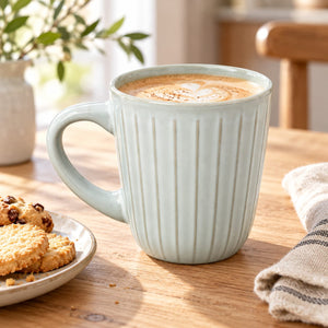 A light sage green ribbed ceramic mug filled with a latte on a wooden table, with cookies on a plate and a striped cloth nearby.