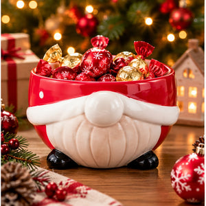 A festive ceramic bowl shaped like a Santa Claus face, filled with red and gold wrapped chocolates, set on a wooden table with Christmas decorations and lights in the background.