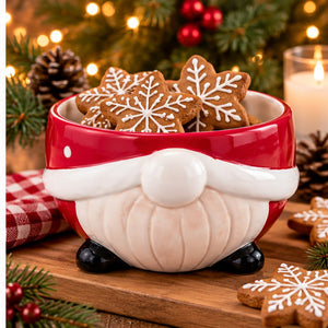 A ceramic Christmas gnome cookie bowl in red and cream, filled with decorated gingerbread cookies, set on a wooden board with holiday lights and pine branches in the background.