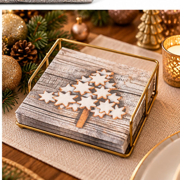 A square gold-framed napkin holder filled with rustic gray wood-patterned napkins, decorated with white iced cookies shaped like a Christmas tree, set on a burlap table runner with holiday decorations in the background.