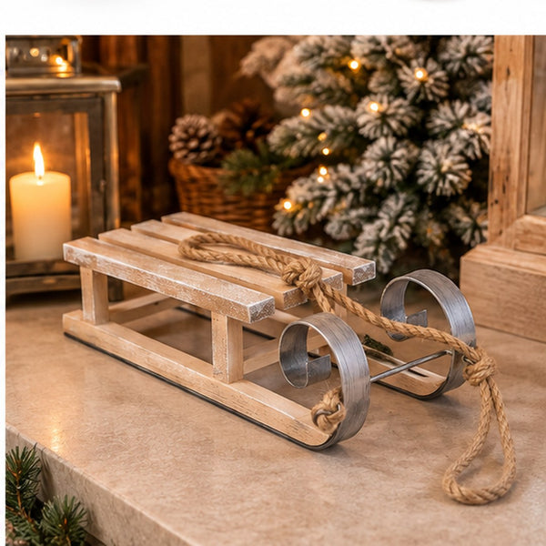 A small wooden sled ornament or decoration with metal wheels and a rope handle, displayed on a tabletop in a warm holiday setting with a candle, lantern, and decorated tree in the background.