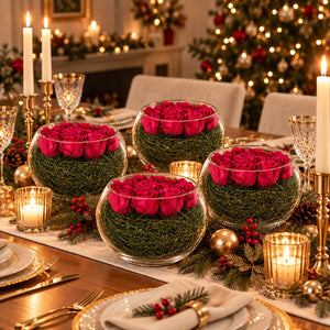 Four round glass bowls filled with green grass and bright pink roses, arranged on a decorated Christmas dinner table with candles, plates, pinecones, and a lit Christmas tree in the background.
