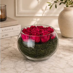 A round clear glass bowl filled with deep pink preserved roses on top of green moss, placed on a light marble surface in a styled interior setting.