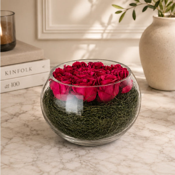 A round clear glass bowl filled with deep pink preserved roses on top of green moss, placed on a light marble surface in a styled interior setting.