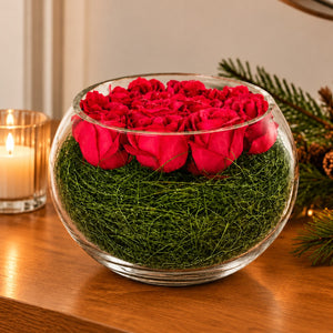 A round clear glass bowl filled with bright red roses on top of green artificial grass, displayed on a wooden table with a lit candle and pine branches in the background.
