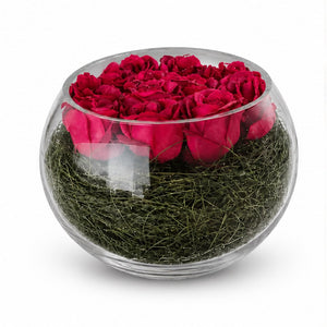 A round clear glass bowl filled with deep red preserved roses on top of green moss, photographed on a clean white background.