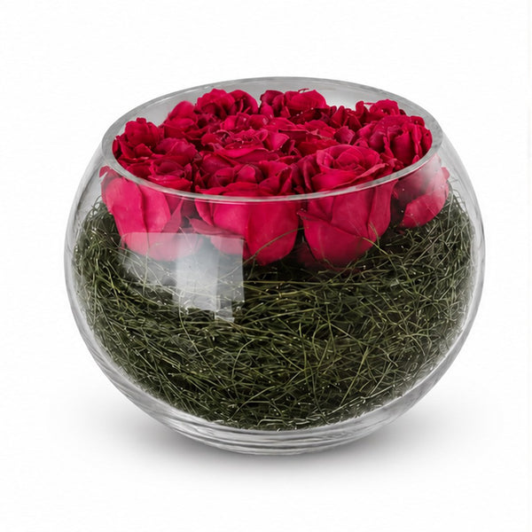A round clear glass bowl filled with deep red preserved roses on top of green moss, photographed on a clean white background.