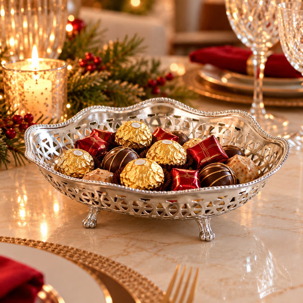 An ornate silver footed bowl filled with assorted chocolates, including Ferrero Rocher, styled on a holiday table with candles, pine branches, and glassware.